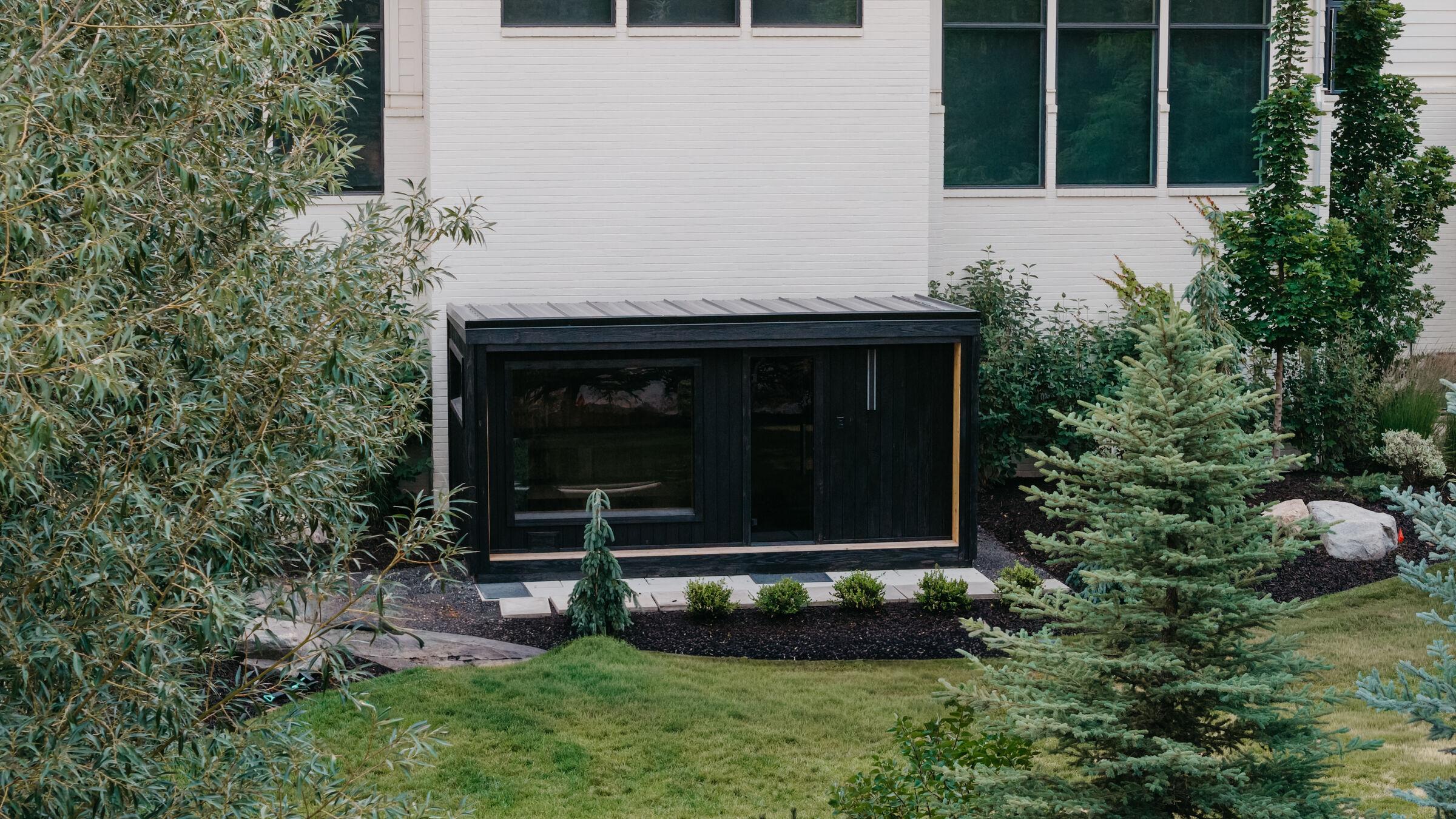 A blackened-timber studio on a mown lawn, framed by young conifers and mulched planting beds